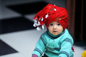 Indian baby girl sitting on a toy horse dressed in green winter sweater and red turban on her...