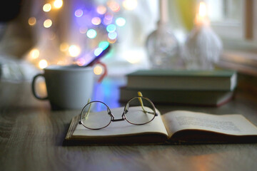 Cup of warm drink with cinnamon stick and candy cane, open book, reading glasses, lit candles and...