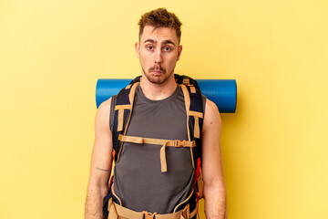 Young hiker caucasian man isolated on yellow background shrugs shoulders and open eyes confused.