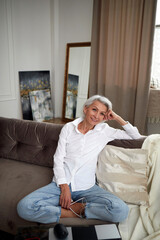 Obraz premium High angle of charming middle aged female with gray hair and in jeans and white shirt sitting with crossed legs on couch at home and smiling at camera