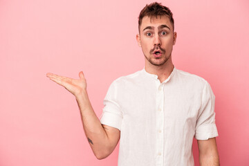 Young caucasian man isolated on pink background impressed holding copy space on palm.