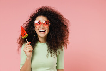 Happy young curly latin woman 20s wears mint t-shirt sunglasses look aside hold eat watermelon...