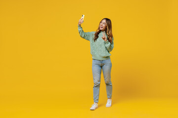 Full body young smiling woman 30s wearing green knitted sweater doing selfie shot on mobile cell phone show v-sign isolated on plain yellow color background studio portrait. People lifestyle concept.