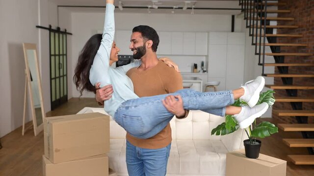 Pakistani husband carrying and embracing wife while feeling delighted, holds her in his arms and whirls, enjoying relocation and starting living together. Home of our own, glad to move into new house