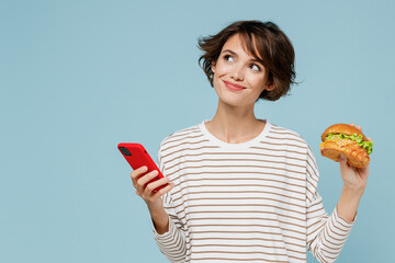 Young minded fun woman in striped shirt using mobile cell phone hold burger count calories browsing internet think isolated on plain pastel light blue background studio People lifestyle food concept