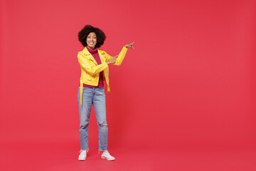 Full size body length happy fancy young curly black latin woman 20s years old wears yellow jacket pointing aside on workspace area copy space mock up isolated on plain red background studio portrait.