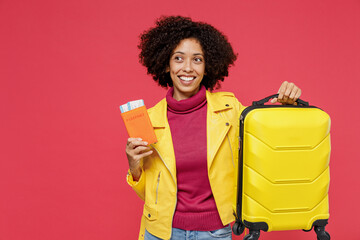 Traveler tourist young curly black latin woman 20s wears yellow jacket hold suitcase bag passport boarding tickets isolated on plain red background studio portrait. Passenger travel abroad getaway