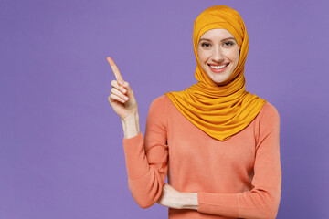 Promoter smiling fun young arabian asian muslim woman in abaya hijab yellow clothes pointing away on workspace area copy space mock up isolated on plain pastel light violet background studio portrait.