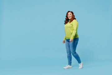 Full body side view happy young chubby overweight plus size big fat fit woman wear green sweater walking going strolling isolated on plain blue background studio portrait. People lifestyle concept
