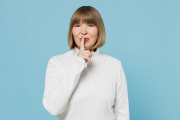 Elderly secret woman 50s wearing white knitted sweater say hush be quiet with finger on lips shhh gesture look camera isolated on plain blue color background studio portrait. People lifestyle concept.