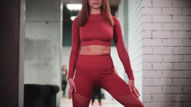 Young woman in red sports suit doing fitness exercises in the gym - looks in the camera and warming up her legs