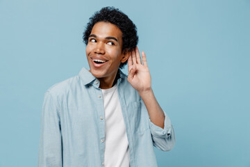 Curious nosy shocked surprised happy young black curly man 20s years old wears white shirt try to hear you overhear listening intently isolated on plain pastel light blue background studio portrait.