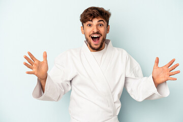 Young mixed race man doing karate isolated on blue background receiving a pleasant surprise,...