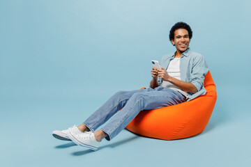 Full size body length fun young black curly man 20s wear white shirt sit in bag chair hold in hand use mobile cell phone typing browsing isolated on plain pastel light blue background studio portrait