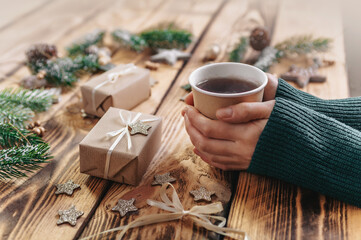 Female hands holding a hot drink on the background of a wooden table with gifts, fir branches and items for creativity