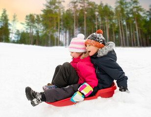 Fototapeta na wymiar childhood, sledging and season concept - happy little kids sliding on sled down snow hill in winter over pine forest on background