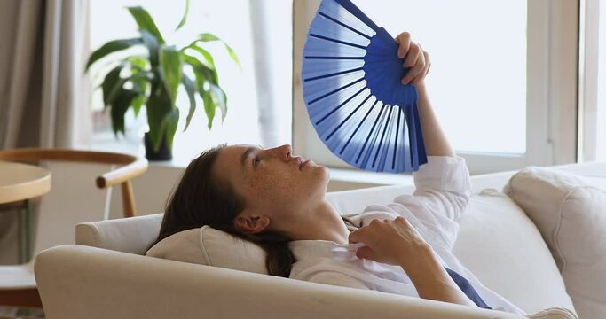 Young freckled woman lean on sofa cushions waving fan, cooling herself at hot day, reduces heat inside living room without air-condition, climate control system. Discomfort, hormonal imbalance concept