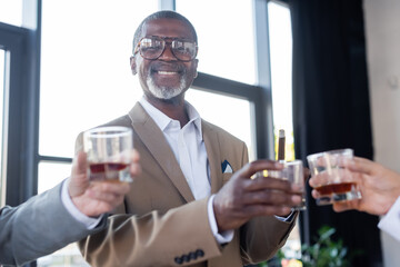 cheerful african american businessman looking at camera near blurred colleagues with glasses of whiskey