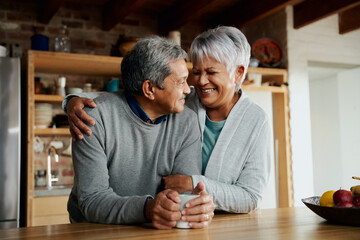 Happily retired elderly biracial couple smiling at each other. Wife holding husband in modern kitchen.  - 474695234