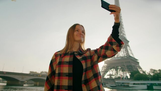 Millennial Woman takes Selfie Photo by Smartphone with Eiffel Tower in Paris, France on Sunny Day. Vacation and Tourism in Europe, Travel Blogger shoots European Landmarks. 4K medium orbit shot