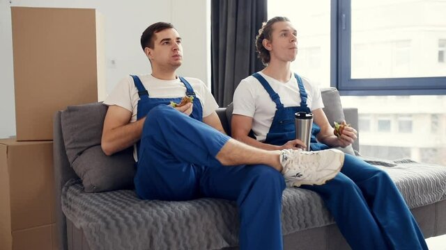 Tired and taking break. Two young movers in blue uniform working indoors in the room