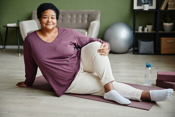 Full length portrait of active senior woman doing yoga at home on floor and looking at camera