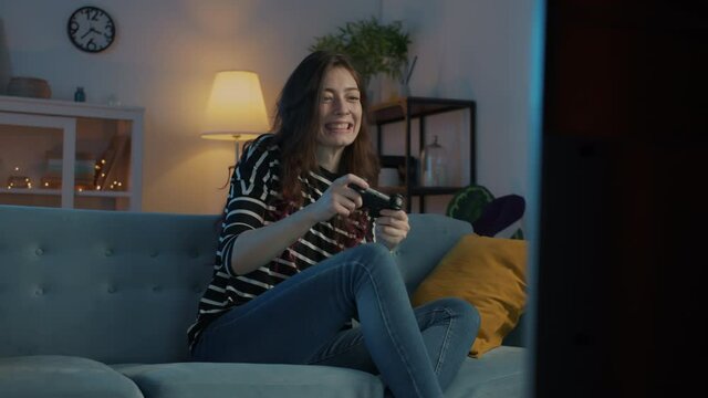 Young pretty girl is enjoying video game at home at night sitting on sofa in modern apartment. Carefree youth and modern lifestyle concept.