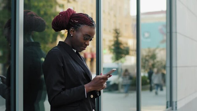 Beautiful young african american girl business woman user model student female lady standing in city on street near building typing message using mobile phone smartphone smiling chatting with device