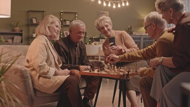 Medium shot of senior friends get together in big cozy living room with lights in evening. Men playing chess while women sitting nearby watching. One of women taking video on smartphone
