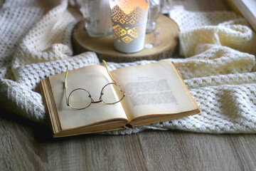 Open book and reading glasses on the table. Lit candles, soft blanket and Christmas decorations in the background. Hygge at home. Selective focus.