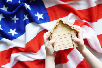 Toy wooden house in a child's hands above the USA flag. 4th July or American citizenship concept.