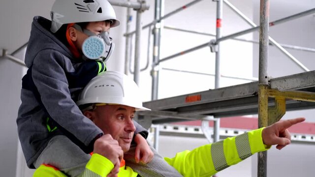 experienced male builder in yellow protective jacket, child, boy in white hardhats boy on shoulders, father, grandfather tells grandchildren about his profession with background of high buildings