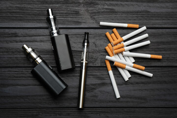 Cigarettes and different vaping devices on black wooden background, flat lay. Smoking alternative