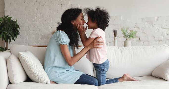 Loving African mother her cute little daughter have fun sit on sofa, smile touch noses, enjoy warmth harmonic relation, moment of tenderness. Happy motherhood, priceless time together, cherish concept