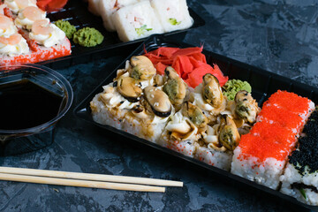 Sushi rolls with mussels, caviar, wasabi, ginger, cheese, shrimp, chicken, soy sauce and cucumber in plastic packaging for takeaway on a dark background
