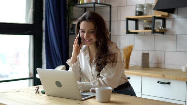 Young female having mobile phone conversation while sitting front open laptop computer at home kitchen interior. Attractive brunette woman calling with smartphone for discuss work issues 