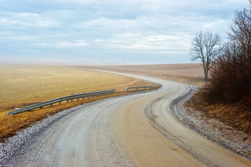 A country dirt road winds and bends towards the horizon of a cloudy and grey winter sky.