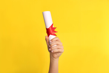 Student holding rolled diploma with red ribbon on yellow background, closeup