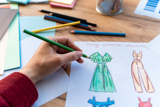 Cropped view of the designer making sketches of clothes while sitting at the table. Drawing, markers, pencil, paper laying at the wooden table. Stock photo