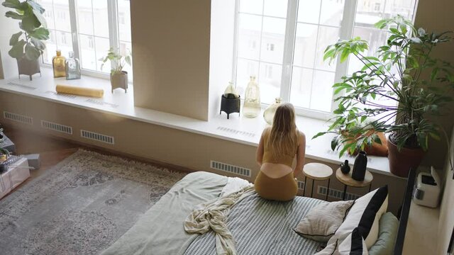 Young woman with blonde long hair wearing yellow sports outfit stretches standing up from soft bed and looking out of window upper view