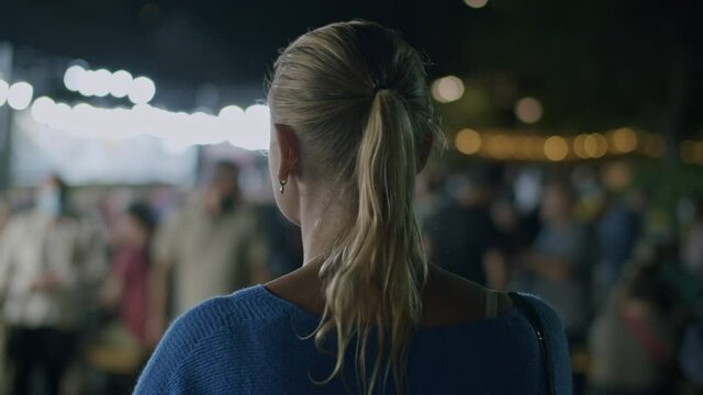 Back view of a blond woman walking in night city and taking pictures with smartphone. Person against blurred crowded street