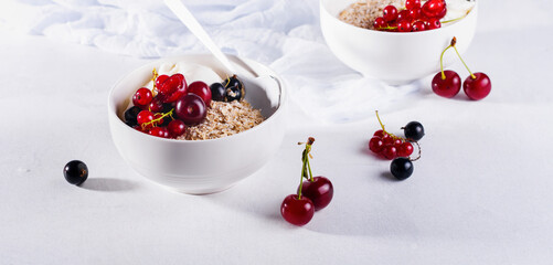 Raw oatmeal with yogurt and berries of cherry, currant and blueberry for making healthy breakfast in white ceramic bowl on a light background. Selective focus.