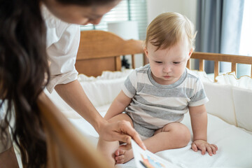 Caucasian mom read storytale to baby infant in child crib in bedroom.