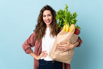 Young woman holding a grocery shopping bag isolated on blue background posing with arms at hip and...