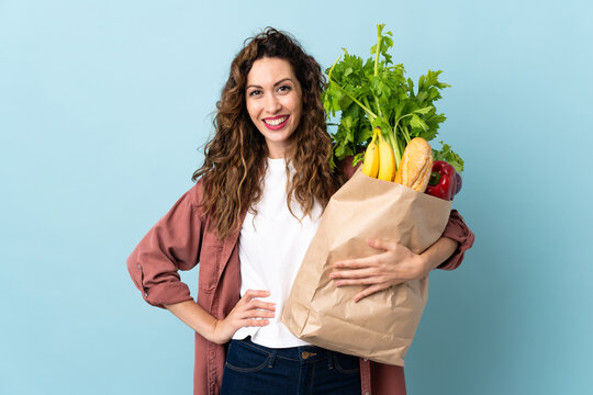 Young woman holding a grocery shopping bag isolated on blue background posing with arms at hip and smiling