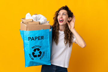 Young woman holding a recycling bag full of paper to recycle isolated on yellow background listening to something by putting hand on the ear