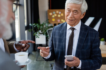 senior asian businessman with smartphone smiling near blurred multiethnic colleagues