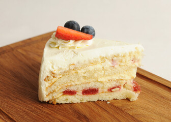 Piece of milk cake on wooden board. Biscuit layers, butter cream, berry jam, cheese glaze. Decorated with fresh berries