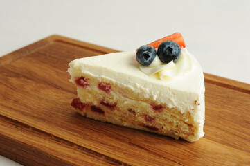 Piece of milk cake on wooden board. Biscuit layers, butter cream, berry jam, cheese glaze. Decorated with fresh berries