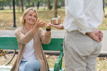 Happy couple senior man surprise giving gift box to his wife while relaxing and sitting on the bench in the park